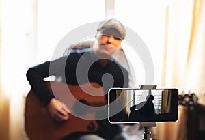 Man with acoustic guitar in a shirt and cap plays live staying home for the audience in front of the smartphone camera during