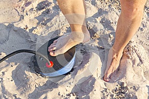 A Man with air foot pump pumps an inflatable mattress or air bed at sandy beach. Foot inflates air mattress with foot