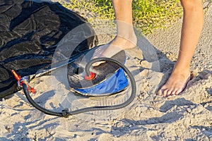 A Man with air foot pump pumps an inflatable mattress or air bed at sandy beach. Foot inflates air mattress with foot