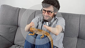 Man with animal bearded agama sitting on sofa at home and stroking reptile.