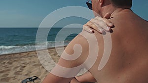 Man is applying sunscreen cream in his skin, sitting on a beach in summer day