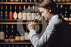 Man appreciating white wine from glass standing in cellar against fulled wooden shelf background with bottles, close up. photo