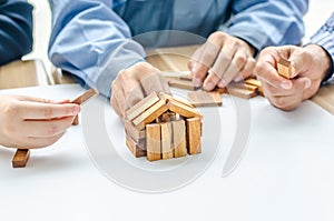 Two man one women architect draws a plan,on large sheet of paper at office desk and builds model house from wooden blocks bars.