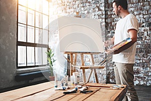 Man artist stands in front of a blank canvas on easel with palette in loft art studio. Male painter hold paintbrush in hand and mi