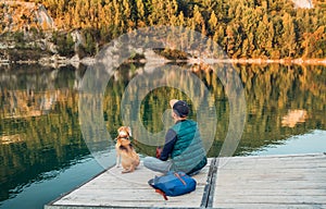 Man as a dog owner and his friend beagle dog are sitting on the wooden pier on the mountain lake and enjoying the landscape during