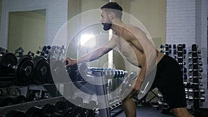 A man of athletic builds exercises his arm muscles with a dumbbell in the gym in front of a mirror next to a racks of