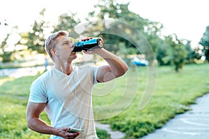 A man atlen drinks water during exercise, with headphones, a smartphone in his hand. In the summer in the city, a