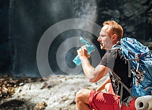 Man with backpack dressed in active trekking clothes drinking the water from touristic flask sitting near mountain river waterfall