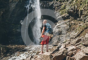 Man with backpack dressed in active trekking clothes holding trekking boots in hand near mountain river waterfall smiling and