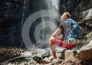 Muž s batohom oblečený v aktívnom trekingovom oblečení si vyzúva trekingové topánky, sedí pri vodopáde horskej rieky a užíva si