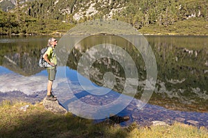 A man with a backpack stands against the backdrop of a mountain lake with a reflected sky, mountain and clouds