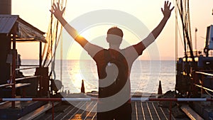 Man with backpack stands on a pier with rising hands during beautiful sunset. Concept of happy travels