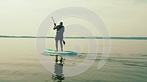 A man with a backpack on a SUP board swims in the lake on a sunny day against a sky with clouds