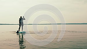 A man with a backpack on a SUP board swims in the lake on a sunny day against a sky with clouds