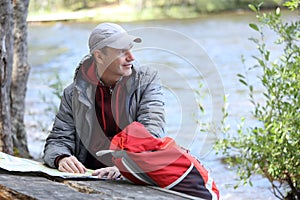 Man with backpack and tourist map sitting against lake Saimaa and looking away