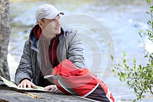 Man with backpack and tourist map sitting against lake Saimaa and looking away