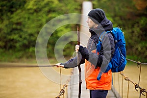 Man with backpack trekking in forest by hinged bridge over river. Cold weathe. Spring hiking. Wooden bridge across the river