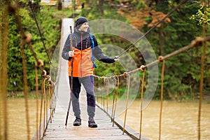 Man with backpack trekking in forest by hinged bridge over river. Cold weathe. Spring hiking. Wooden bridge across the river