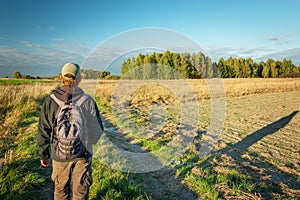 A man with a backpack walking dirt road through fields, forest and blue sky