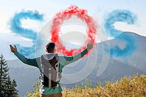 Man with backpack and word SOS made of color smoke bomb in mountains