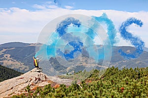 Man with backpack and word SOS made of smoke bomb on rocky peak in mountains, back view