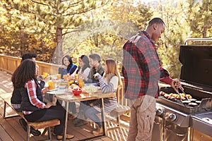 Man barbecues for friends at a table, on a deck in a forest