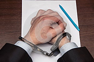 A man with bare hands in handcuffs sits at a table in front of a blank sheet of paper and a fountain pen. Concept: the detainee