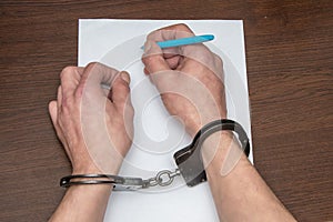 A man with bare hands in handcuffs sits at a table in front of a blank sheet of paper and a fountain pen.