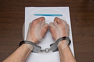 A man with bare hands in handcuffs sits at a table in front of a blank sheet of paper and a fountain pen. 2