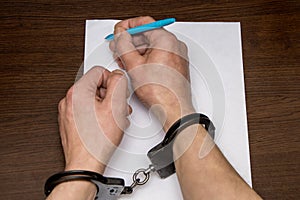 A man with bare hands in handcuffs sits at a table in front of a blank sheet of paper and a fountain pen. 7