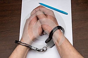 A man with bare hands in handcuffs sits at a table in front of a blank sheet of paper and a fountain pen. 3