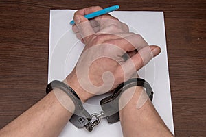 A man with bare hands in handcuffs sits at a table in front of a blank sheet of paper and a fountain pen. 6