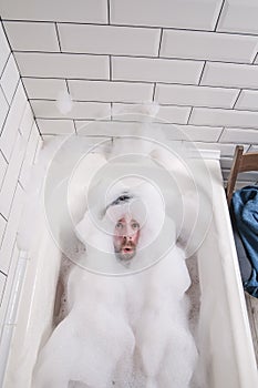 Man is bathing in a bathtub, he is covered in lush soap suds, only his face with a funny expression is visible.