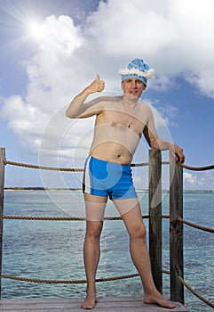 Man in a bathing suit and New Year Santa Klaus cap shows a thumb on sea background