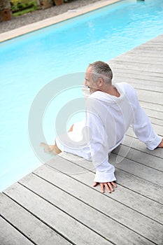 Man in bathrobe sitting on wood deck of pool