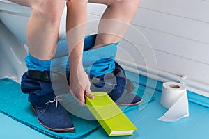 man in the bathroom, sitting on the toilet, with his pants down and holding a book