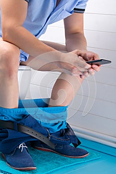 A man in the bathroom, sitting on the toilet, with his pants down and holding a mobile phone