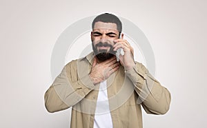 Man with a beard appears distressed while talking on the phone, hand on throat suggesting difficulty speaking