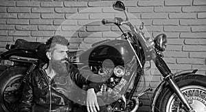 Man with beard, biker in leather jacket near motor bike in garage, brick wall background. Bikers lifestyle concept