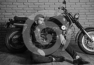 Man with beard, biker in leather jacket near motor bike in garage, brick wall background. Hipster, brutal biker on