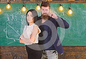 Man with beard and girl teacher stand in classroom, chalkboard on background. Teacher and strict schoolmaster look