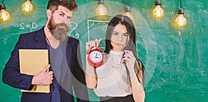 Man with beard hold book and girl teacher holds alarm clock, chalkboard on background. School rules concept. Lady