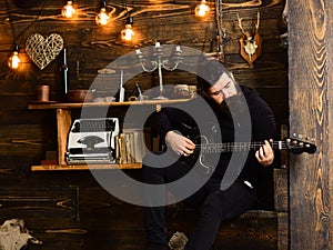 Man with beard holds black electric guitar. Guy in cozy warm atmosphere play music. Man bearded musician enjoy evening