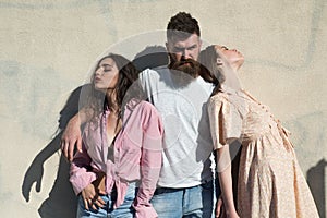 Man with beard hugs two ladies on hot sunny day. Girls turned to opposite sides while man hugs them. Love triangle