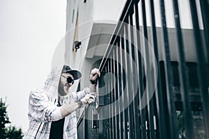 Man with beard in light checkered jacket with hood in a cap and sunglasses picks up the apartment number on the keypad to call the