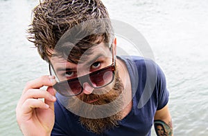 Man with beard and mustache peeking out of sunglasses, river on background. Hipster on confident face with fashionable