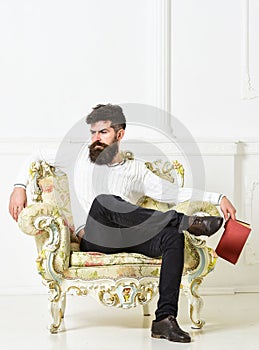 Man with beard and mustache sits on armchair, holds book, white wall background. Connoisseur on thoughtful face finished photo