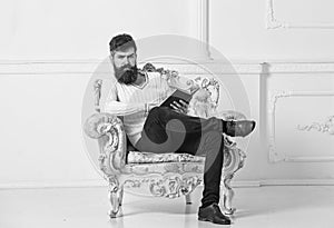 Man with beard and mustache sits on armchair and reading, white wall background. Connoisseur, professor enjoy literature