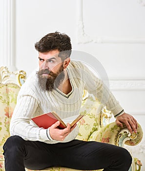 Man with beard and mustache sits on baroque style sofa, holds book, white wall background. Reflections on literature