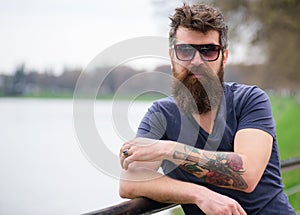 Man with beard and mustache wears sunglasses, river embankment on background. Hipster on confident face with fashionable
